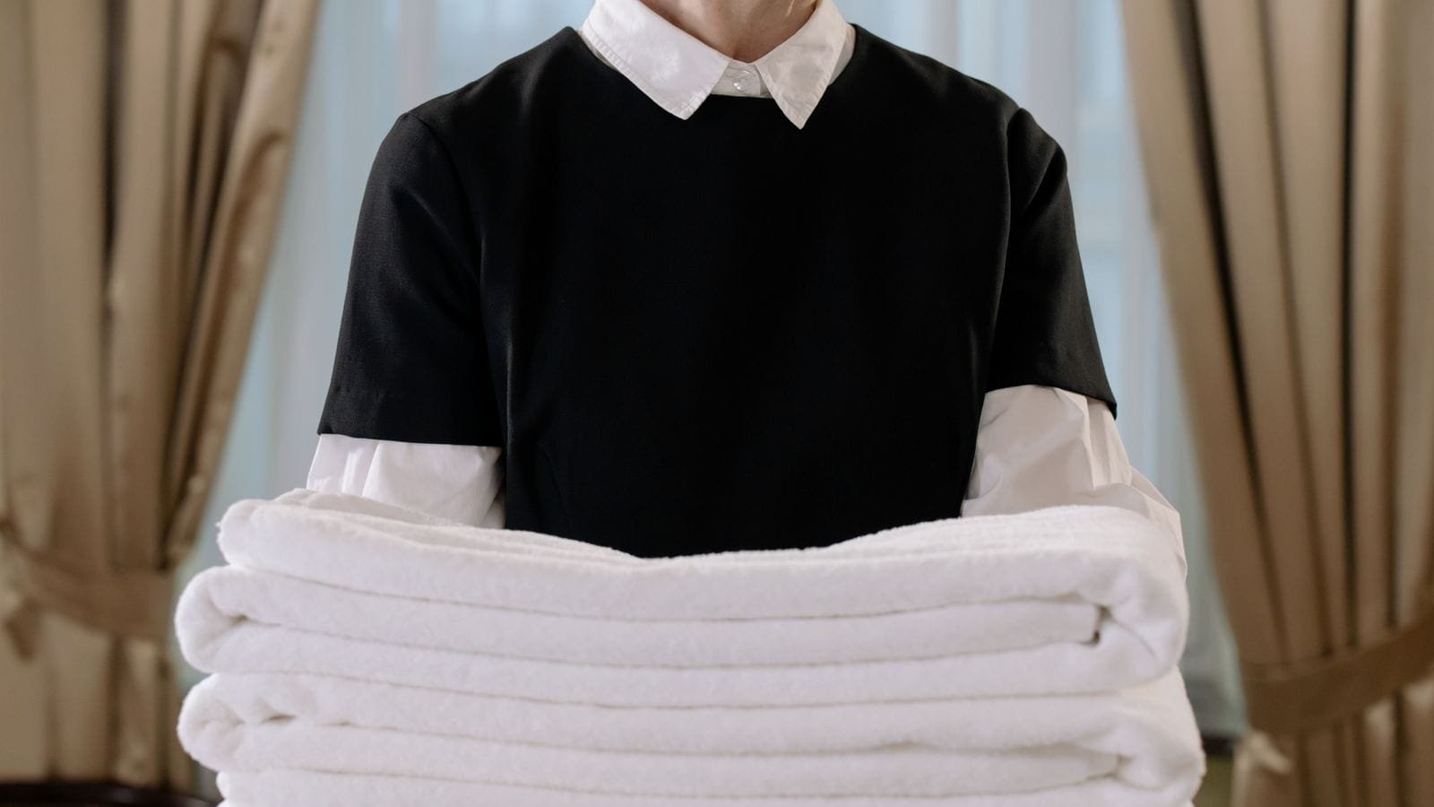 Camareira de hotel em uniforme preto segurando uma pilha de toalhas brancas dobradas